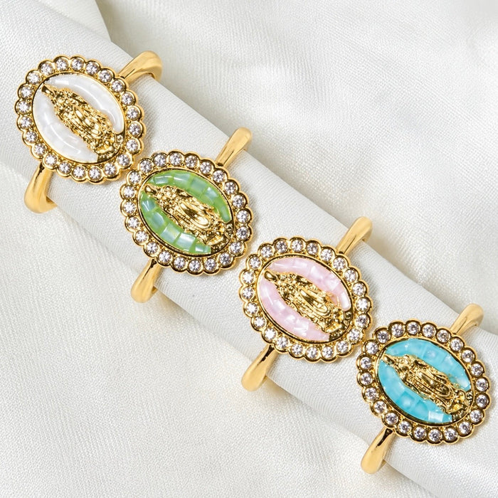 Four gold-plated rings with oval-shaped resin inserts featuring religious figures surrounded by clear rhinestones, arranged on a white satin fabric. The inserts display different pastel colors: white, light green, light pink, and light blue.