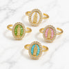 Four gold-toned adjustable rings with oval Miraculous Medal enameled faces, arranged on a white marble background. The medallions feature white, green, pink, and turquoise backgrounds.