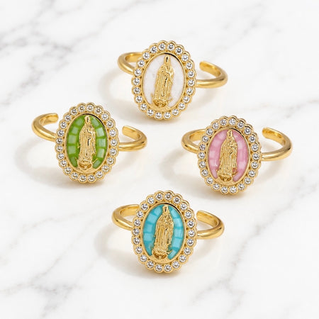 Four gold-toned adjustable rings with oval Miraculous Medal enameled faces, arranged on a white marble background. The medallions feature white, green, pink, and turquoise backgrounds.