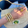 A close-up of a hand wearing multiple gold-toned jewelry pieces, including a large floral ring, a stacked bangle set with small red and white stones, and a silver-toned band, resting on a woven straw handbag.