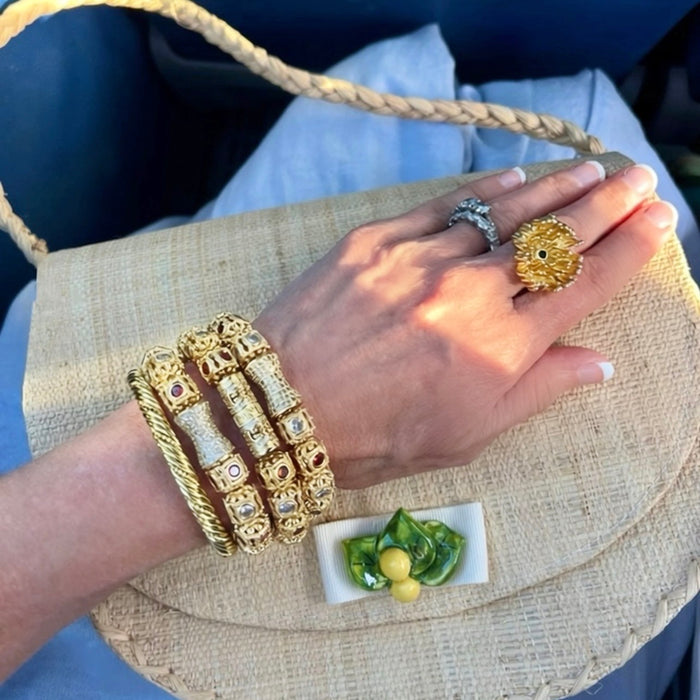 A close-up of a hand wearing multiple gold-toned jewelry pieces, including a large floral ring, a stacked bangle set with small red and white stones, and a silver-toned band, resting on a woven straw handbag.
