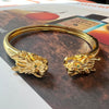 A gold-toned open cuff mahjong bracelet with detailed dragon head motifs at both ends, photographed on top of printed magazine pages.