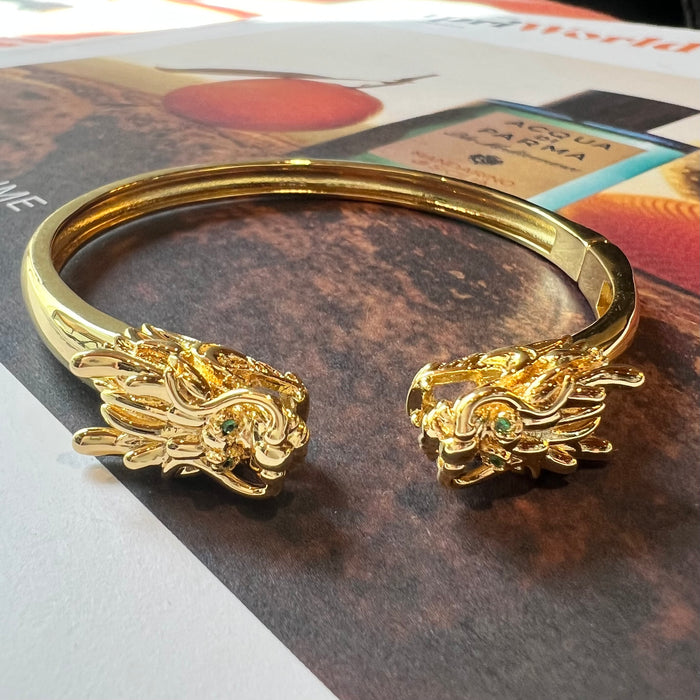 A gold-toned open cuff mahjong bracelet with detailed dragon head motifs at both ends, photographed on top of printed magazine pages.