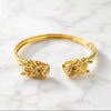 A gold-toned open cuff mahjong bracelet with dragon head motifs at both ends, resting on a white marble surface. 