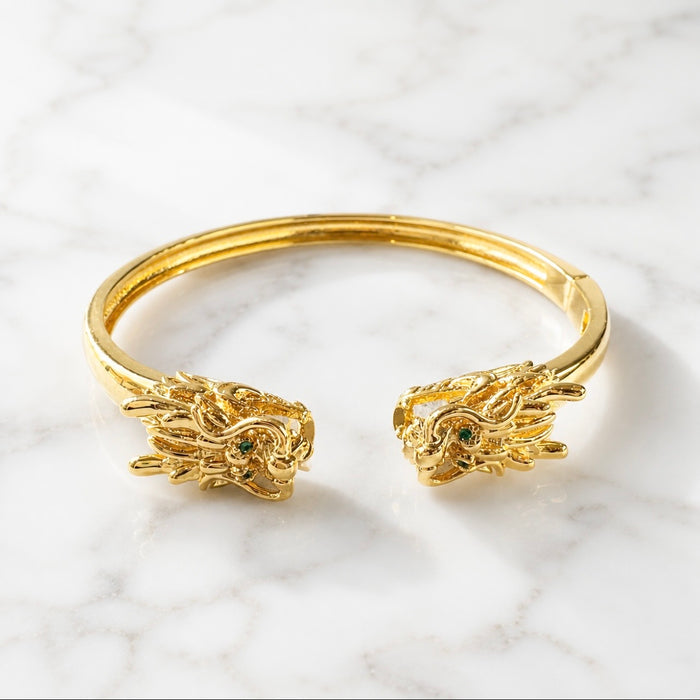 A gold-toned open cuff mahjong bracelet with dragon head motifs at both ends, resting on a white marble surface. 