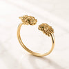 A gold open cuff ring featuring two sculpted mahjong dragon head ends with small green gemstone eyes, displayed on a light marble surface.