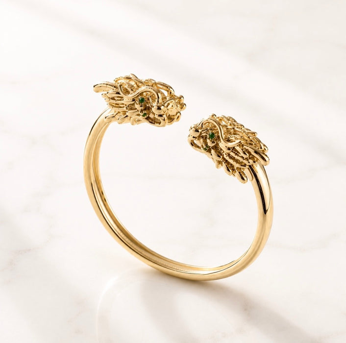 A gold open cuff ring featuring two sculpted mahjong dragon head ends with small green gemstone eyes, displayed on a light marble surface.