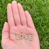 A close-up of a person's open hand holding three gold letter pendants spelling LOVE, each set with small clear stones, against a blurred green grass background.