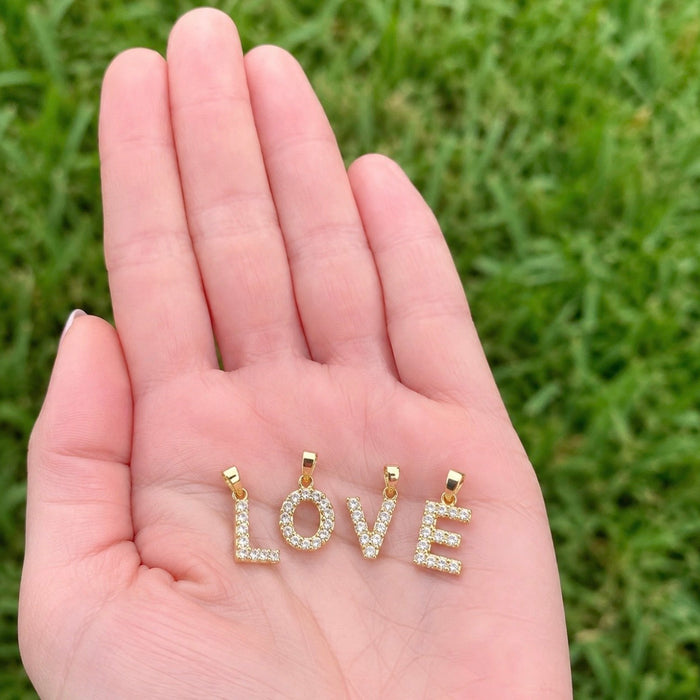 A close-up of a person's open hand holding three gold letter pendants spelling LOVE, each set with small clear stones, against a blurred green grass background.