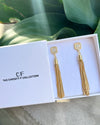 Gold tassel earrings in a 'The Christy F Collection' box with green leaves in the background.
