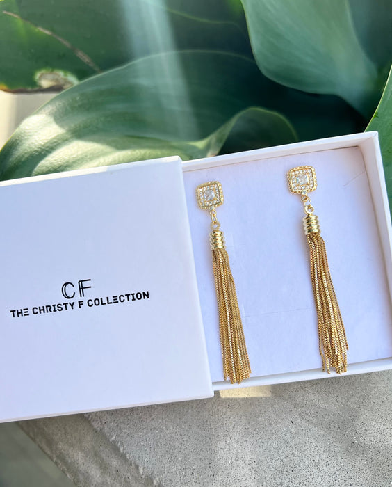 Gold tassel earrings in a 'The Christy F Collection' box with green leaves in the background.