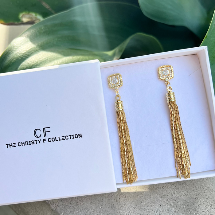 Gold tassel earrings in a 'The Christy F Collection' box with green leaves in the background.