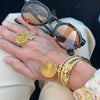A close-up of a person's hand wearing multiple gold jewelry pieces, including a large textured gold ring, a pink stone ring, and a gold bangle with a round sunburst charm, alongside tortoiseshell sunglasses resting on the hand.