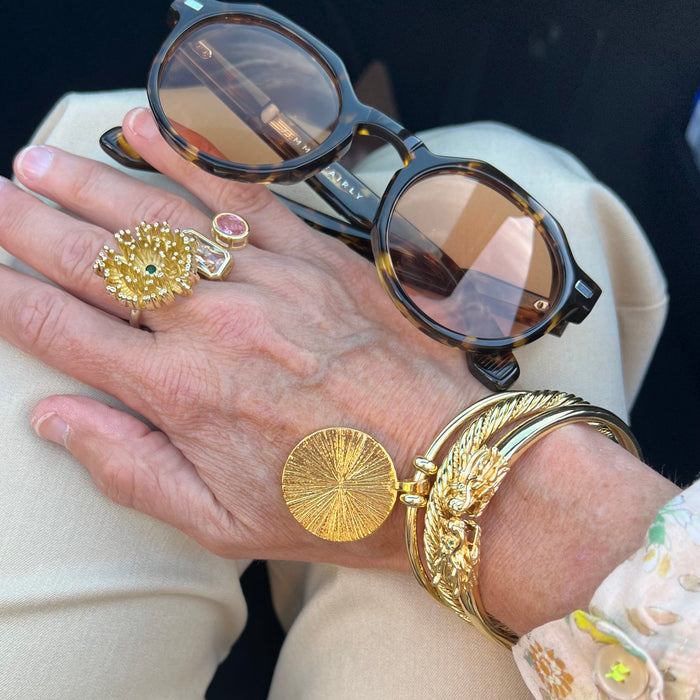 A close-up of a person's hand wearing multiple gold jewelry pieces, including a large textured gold ring, a pink stone ring, and a gold bangle with a round sunburst charm, alongside tortoiseshell sunglasses resting on the hand.