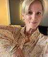 A close-up selfie of a woman with short blonde hair wearing a beige floral blouse with puff sleeves, gold necklaces, and a gold bracelet.