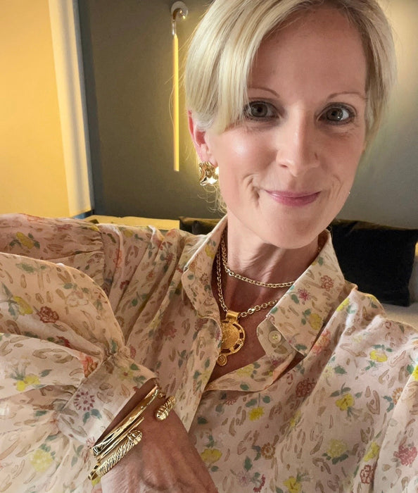 A close-up selfie of a woman with short blonde hair wearing a beige floral blouse with puff sleeves, gold necklaces, and a gold bracelet.