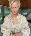 A blonde woman wearing a cream-colored floral button-up blouse with gold jewelry, including a large gold pendant necklace, gold earrings, and gold bracelets, standing in a modern indoor setting.