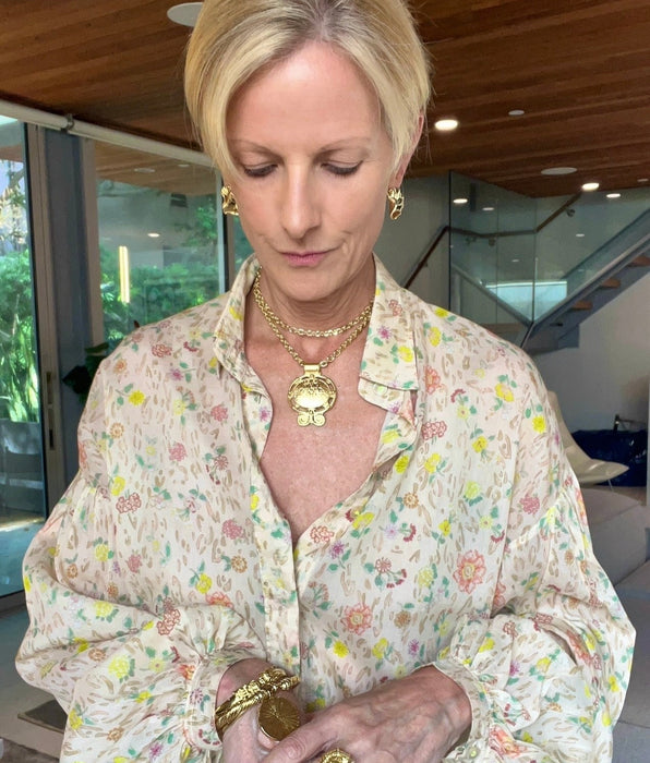 A blonde woman wearing a cream-colored floral button-up blouse with gold jewelry, including a large gold pendant necklace, gold earrings, and gold bracelets, standing in a modern indoor setting.