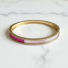 A gold bangle bracelet featuring a row of baguette-cut gemstones in pink and magenta, displayed on a white marble surface.