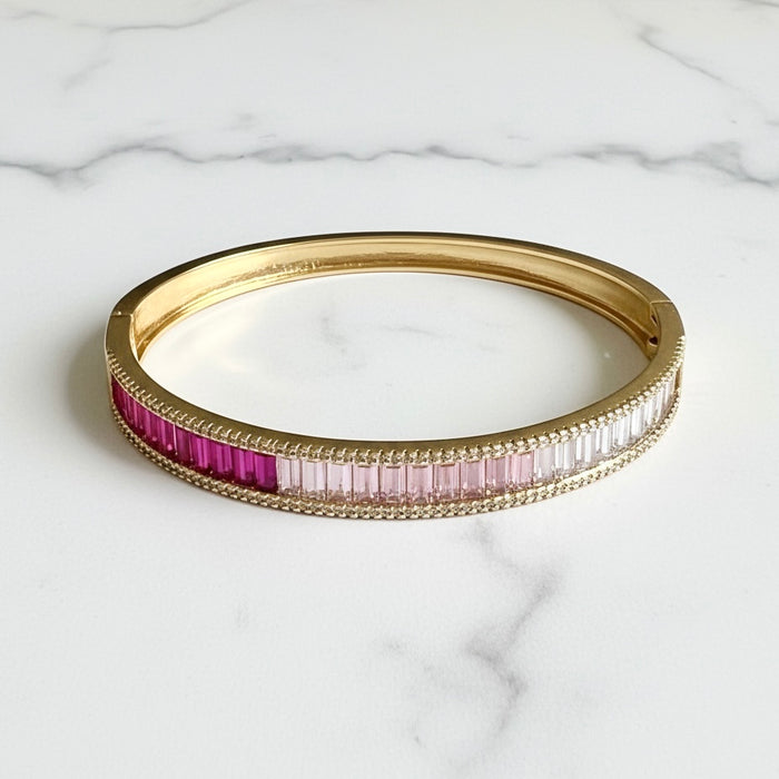 A gold bangle bracelet featuring a row of baguette-cut gemstones in pink and magenta, displayed on a white marble surface.