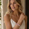 A woman with blonde hair wearing a white spaghetti-strap camisole top and a white beaded bracelet with pink beads, shown in a warm, sunlit indoor setting.