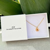 A gold necklace with a circular pendant featuring a red eye design, presented in a white jewelry box with the brand name 'The Christy F Collection' printed on the lid. The box is styled on a light gray surface with a green leaf in the background.