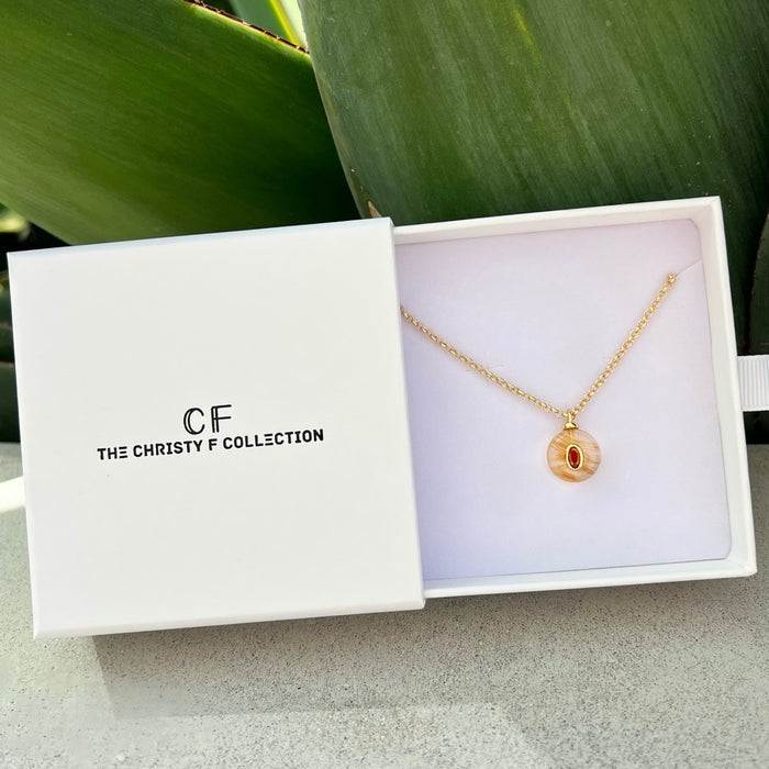A gold necklace with a circular pendant featuring a red eye design, presented in a white jewelry box with the brand name 'The Christy F Collection' printed on the lid. The box is styled on a light gray surface with a green leaf in the background.
