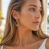 A close-up of a woman wearing a gold hoop Mahjong White Dragon earring with a central oval charm featuring a blue and white pattern, set against a softly blurred outdoor background.