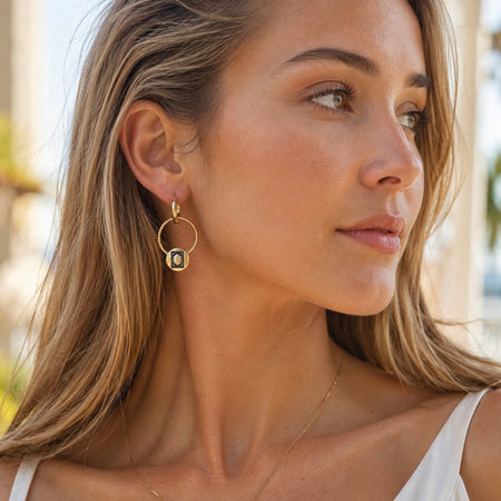 A close-up of a woman wearing a gold hoop Mahjong White Dragon earring with a central oval charm featuring a blue and white pattern, set against a softly blurred outdoor background.