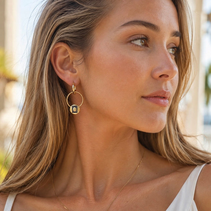 A close-up of a woman wearing a gold hoop Mahjong White Dragon earring with a central oval charm featuring a blue and white pattern, set against a softly blurred outdoor background.