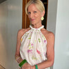 A woman wearing a white halter top with a pink and green floral print, white pants, and holding a green leather handbag, standing indoors wearing gold stud earrings, a green bangle, gold bangle and lavender bangle with three gold rings.