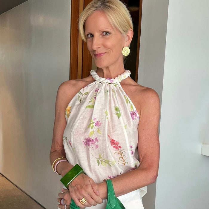 A woman wearing a white halter top with a pink and green floral print, white pants, and holding a green leather handbag, standing indoors with gold earrings, three gold rings, purple bracelet and gold bangle.