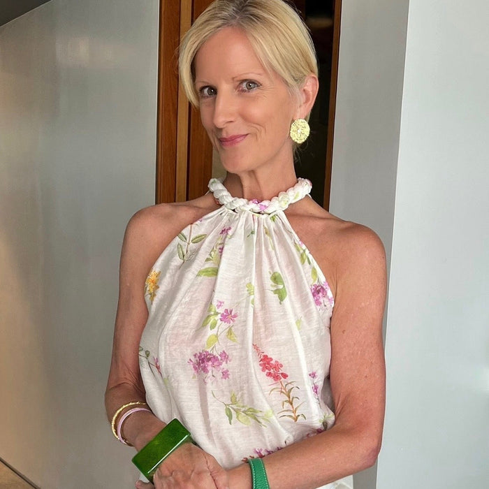 A woman wearing a white halter top with a pink and green floral print, white pants, and holding a green leather handbag, standing indoors wearing gold stud earrings, a green bangle, gold bangle and lavender bangle with three gold rings.