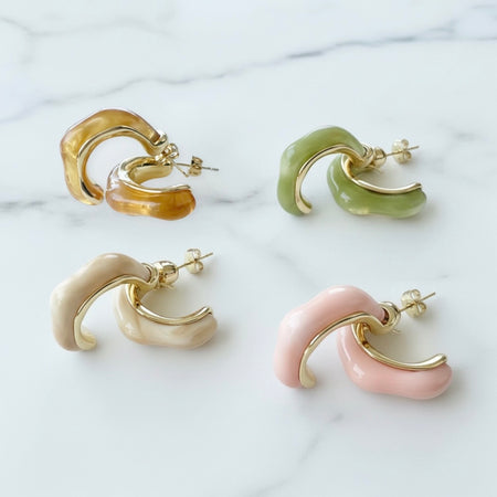 Four curved hoop-style earrings arranged on a white marble surface, shown in amber, light green, cream, and blush pink with gold-tone metal edges and posts.