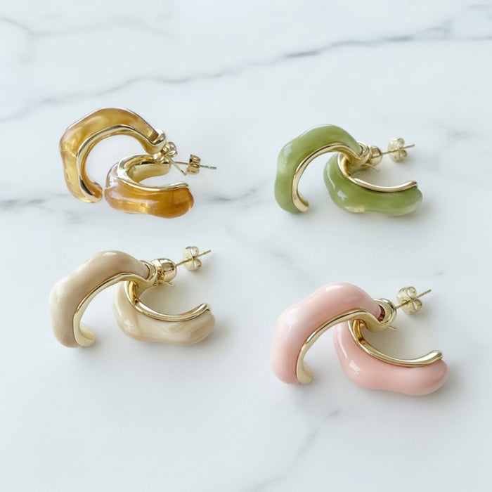 Four curved hoop-style earrings arranged on a white marble surface, shown in amber, light green, cream, and blush pink with gold-tone metal edges and posts.