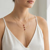 A close-up of a model wearing a delicate gold lariat necklace with three small rectangular red gemstones and a small gold heart pendant, styled with a second thin gold chain necklace.