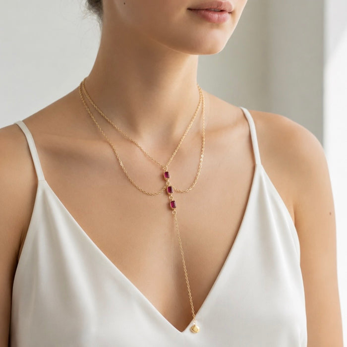 A close-up of a model wearing a delicate gold lariat necklace with three small rectangular red gemstones and a small gold heart pendant, styled with a second thin gold chain necklace.