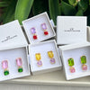 Four pairs of colorful gemstone stud earrings displayed in white jewelry boxes. The earrings feature rectangular and square-cut stones in pink, purple, yellow, green, and red, set in gold-tone metal.