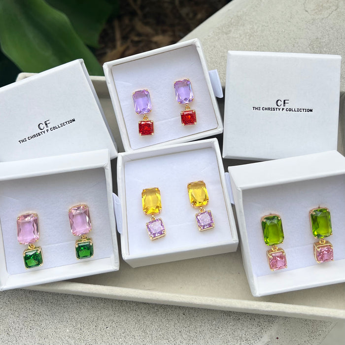 Four pairs of drop earrings displayed in white jewelry boxes on a tray. The earrings feature rectangular gemstones in pink, purple, yellow, green, and red, set in gold-tone metal.