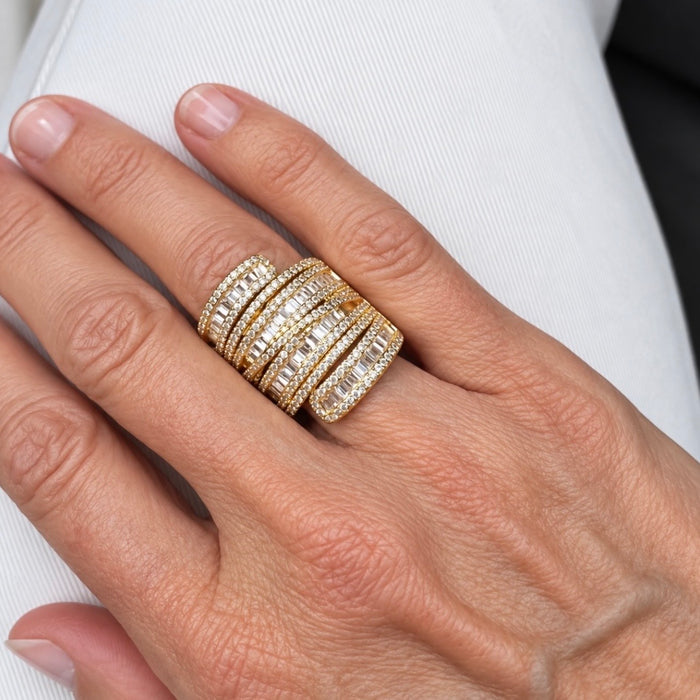 A close-up of a hand wearing a stack of three gold rings set with clear baguette and round stones, resting on white fabric.