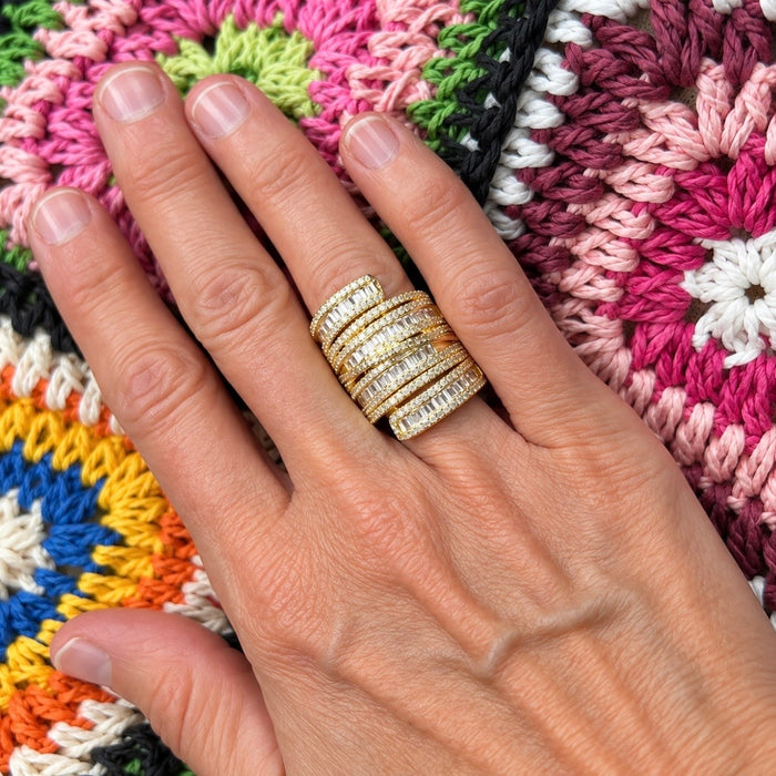 A close-up of a hand wearing a stack of wide gold rings set with clear stones, resting on a colorful crocheted blanket.