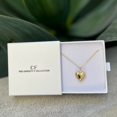 A gold-filled heart-shaped puffy necklace presented in an open white box with 'CF THE CHRISTY F COLLECTION' printed on it, set against a backdrop of a green plant.