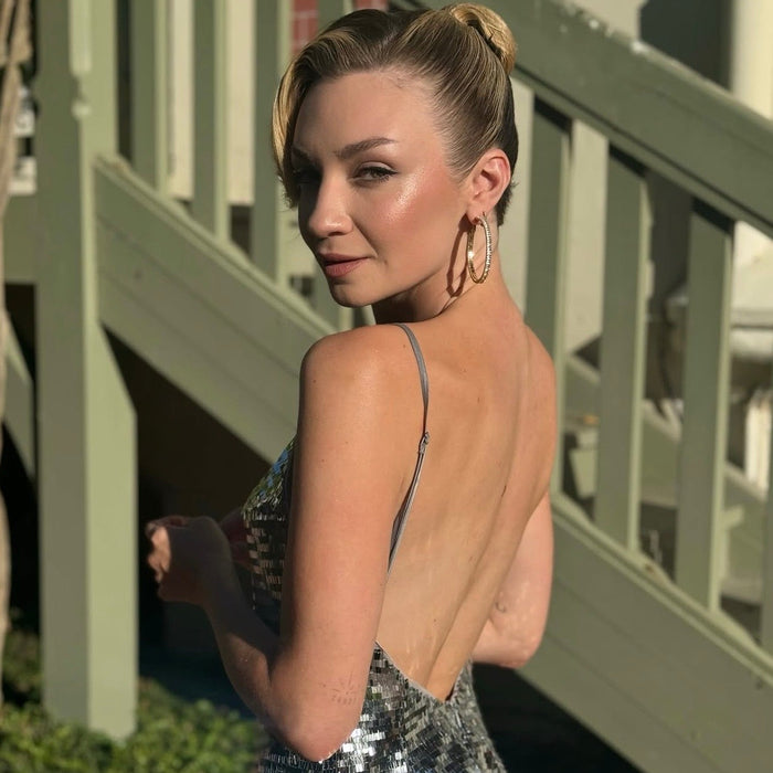 A woman wearing a shimmering silver sequined backless dress and large gold hoop earrings, photographed outdoors against a light green wooden staircase.