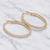 A pair of gold hoop earrings with a textured outer band and a row of clear stones on the inner band, displayed on a white marble surface.