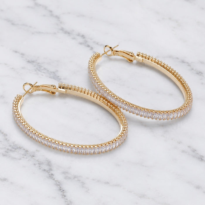 A pair of gold hoop earrings with a textured outer band and a row of clear stones on the inner band, displayed on a white marble surface.