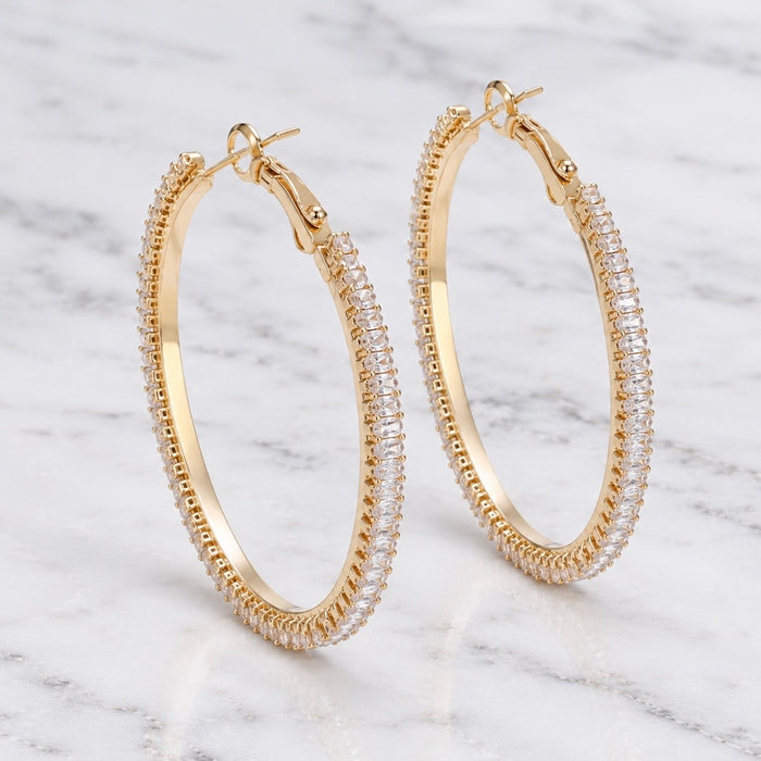 A pair of gold hoop earrings paved with clear cubic zirconia stones, displayed on a white marble surface.