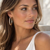 A close-up of a woman wearing large, textured gold hoop earrings, shown from the chest up against a softly blurred light gray background.