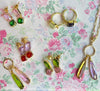 A styled flat-lay of gold jewelry featuring colorful gemstone accents, including hoop earrings, drop earrings, and a necklace, arranged on a pink and green floral patterned background.
