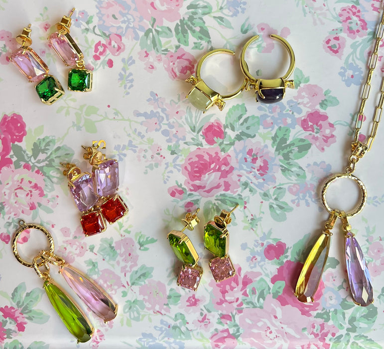 A styled flat-lay of gold jewelry featuring colorful gemstone accents, including hoop earrings, drop earrings, and a necklace, arranged on a pink and green floral patterned background.
