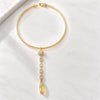 A gold Y-necklace featuring a textured chain, a central round bezel-set clear stone, and a dangling teardrop-shaped yellow gemstone, displayed on a white marble surface.